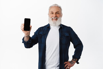 Smiling old man with tattoos shows mobile phone blank screen, looking happy and satisfied, recommending smartphone app, standing over white background