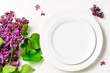 Empty white plate and fresh lilac flowers on white concrete background top view. Spring restaurant 