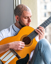Pensive Male With Tattooed Bald Head In Casual Clothes Sitting On Windowsill And Playing Guitar In Daytime
