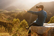 © Alberto Menendez/ADDICTIVE STOCK - Side view of male traveler in VR glasses interacting with virtual reality while sitting on hill in mountainous terrain at sunset