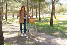 Full Body Of Young Female Standing And Concentrated Near Old Bicycle With Timber Wicker Basket In The Park