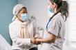 © Alvaro Sanchez/ADDICTIVE STOCK - Young female doctor in medical uniform and stethoscope wearing face mask speaking and holding hands of African American mature woman patient during appointment in clinic