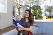 © Christian - Happy mother holding her cute baby with blurry trees and houses as background