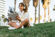 © David Munoz/ADDICTIVE STOCK - Low angle of glad African American female in headphones sitting in exotic park and reading interesting book in summer