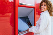 © David Munoz/ADDICTIVE STOCK - Side view of smiling African American female using ATM terminal and withdrawing cash while standing on city street looking at camera