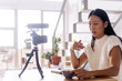 © Jake Jakab/ADDICTIVE STOCK - Young ethnic female vlogger with notebook sitting at table with photo camera on tripod in kitchen