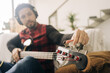 © Jake Jakab/ADDICTIVE STOCK - Bearded male musician in headset tuning bass guitar while sitting on couch in living room