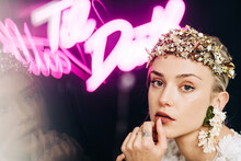 Charming Tender Young Bride In White Lace Gown And Luxurious Floral Wreath And Earrings Looking At Camera Against Black Background With Neon Lights