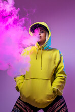 From below confident female hipster in hoodie smoking e cigarette in studio on pink background looking at camera
