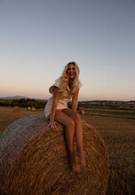 Cheerful Female In Elegant Dress Sitting On Haystack In Dry Field In Rural Area And Looking At Camera