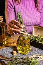 Crop Faceless Female In Purple Sweater Putting Herbs Twigs With Green Leaves In Essential Oil Glass Bottle Near Scissors And Rope With Small Chest On Cloth At Wooden Table