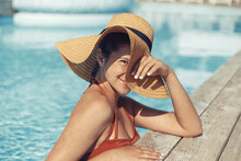 Enjoying summer vacation. Beautiful happy woman in hat relaxing in pool. Portrait of smiling carefree young female sunbathing and resting at swimming pool edge at tropical resort. Travel