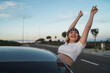 © Miguel Angel Junquera/ADDICTIVE STOCK - Young female in casual wear and headband with American flag print leaning out of car window and raising hands while enjoying freedom during road trip at sunset