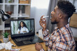 © sofiko14 - Afro american guy using spray for throat to relieve pain while having video call on laptop with female doctor. Concept of people, technology and health care.