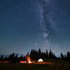 Wall Mural - male traveler looking at campfire while sitting near camp tent under beautiful night sky with stars 