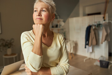 Mature people and aging concept. Indoor shot of bored thoughtful retired female in casual clothing looking away with pensive facial expression holding hand under her chin, wants to go out