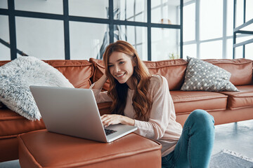 Attractive young woman using laptop and smiling while spending time at home