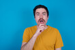 © Roquillo - Nervous puzzled young handsome Caucasian man with moustache wearing orange t-shirt against blue background opens mouth from surprise, reacts on sudden news.