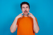 © Roquillo - young handsome Caucasian man with moustache wearing orange t-shirt against blue background crosses eyes and makes fish lips funny grimace