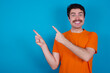 © Roquillo - young handsome Caucasian man with moustache wearing orange t-shirt against blue background points at copy space indicates for advertising gives right direction