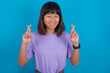 © Roquillo - Young beautiful asian girl wearing purple t-shirt against blue background has big hope, crosses fingers, believes in good fortune, smiles broadly. People and wish concept