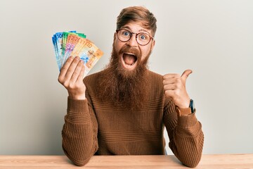 Sticker - Young irish redhead man holding swiss franc banknotes sitting on the table pointing thumb up to the side smiling happy with open mouth