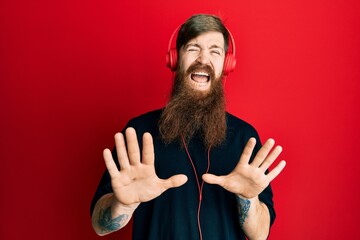 Sticker - Redhead man with long beard listening to music using headphones afraid and terrified with fear expression stop gesture with hands, shouting in shock. panic concept.