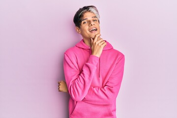 Sticker - Young hispanic man wearing casual pink sweatshirt smiling looking confident at the camera with crossed arms and hand on chin. thinking positive.