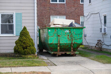 The End Of An Old Blue Green Dumpster Filled With Construction Debris In A Driveway Between Two Houses