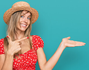 Poster - Beautiful caucasian woman wearing summer hat amazed and smiling to the camera while presenting with hand and pointing with finger.