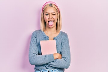 Sticker - Beautiful blonde woman holding book sticking tongue out happy with funny expression.