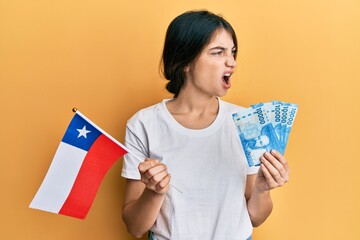 Sticker - Young caucasian woman holding chile flag and chilean pesos banknotes angry and mad screaming frustrated and furious, shouting with anger. rage and aggressive concept.