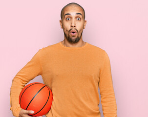 Poster - Hispanic adult man holding basketball ball scared and amazed with open mouth for surprise, disbelief face