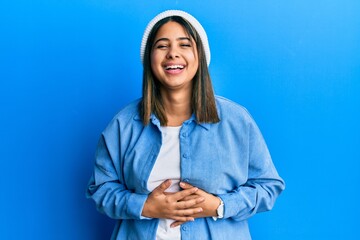 Poster - Young latin woman wearing cute wool cap smiling and laughing hard out loud because funny crazy joke with hands on body.