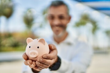 Middle age grey-haired man smiling happy holding piggy bank at the city.