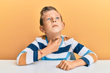 Adorable caucasian kid wearing casual clothes sitting on the table touching painful neck, sore throat for flu, clod and infection