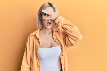 Poster - Young blonde girl wearing casual clothes peeking in shock covering face and eyes with hand, looking through fingers with embarrassed expression.