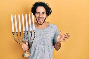 Sticker - Handsome hispanic man holding menorah hanukkah jewish candle celebrating achievement with happy smile and winner expression with raised hand