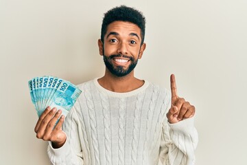 Wall Mural - Handsome hispanic man with beard holding 100 brazilian real banknotes smiling with an idea or question pointing finger with happy face, number one