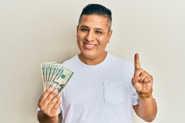 Canvas Print - Young latin man holding 20 dollars banknotes smiling with an idea or question pointing finger with happy face, number one