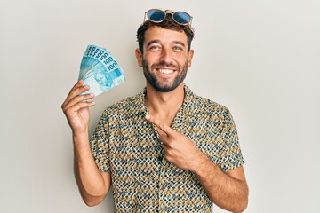 Wall Mural - Handsome man with beard holding 100 brazilian real banknotes smiling happy pointing with hand and finger