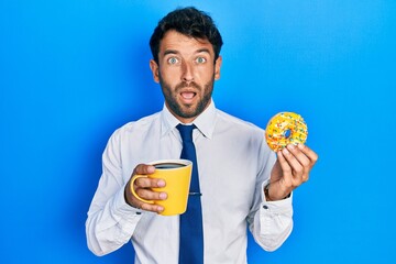 Sticker - Handsome business man with beard eating doughnut and drinking coffee in shock face, looking skeptical and sarcastic, surprised with open mouth