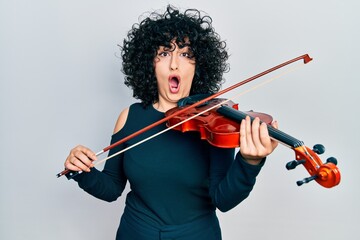 Poster - Young middle east woman playing violin afraid and shocked with surprise and amazed expression, fear and excited face.