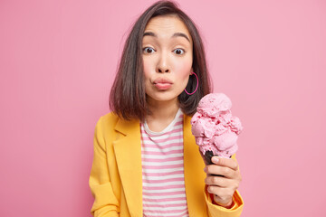 Wall Mural - Pretty millennial Asian woman keeps lips folded holds appetizing ice cream in black waffle spends day off walking outside enjoys summer time eats tasty frozen cold dessert wears yellow jacket