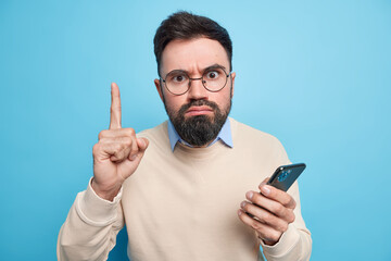 Wall Mural - Strict serious bearded adult man raises index finger has excellent idea uses new mobile application holds smartphone wears spectacles and sweater isolated over blue backgound. Technology user