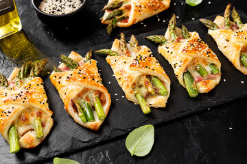 Baked green asparagus with ham and cheese in puff pastry sprinkled with sesame seeds and green basil leaves. Black stone background