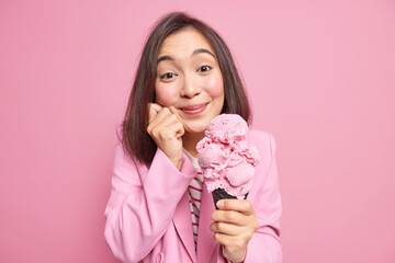 Wall Mural - Attractive brunette young Asian woman with delighted expression holds tasty cone ice cream in waffle keeps hand on face has sweet tooth isolated over pink studio background. Yummy summer dessert