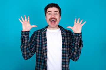 Wall Mural - young Caucasian man with moustache wearing plaid shirt against blue wall raising hands up, having eyes full of happiness rejoicing his great achievements. Achievement, success concept.