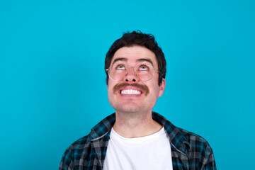 Wall Mural - Portrait of mysterious young Caucasian man with moustache wearing plaid shirt against blue wall looking up with enigmatic smile. Advertisement concept.