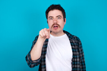 Poster - Shocked young Caucasian man with moustache wearing plaid shirt against blue wall points front with index finger at camera and. Surprise and advertisement concept.